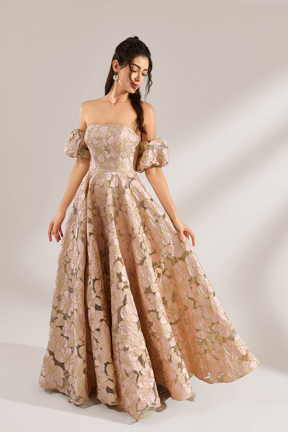 A woman with long, wavy dark hair and a poised stance wears a light - pink, floral - embroidered off - the - shoulder gown. It has puffed sleeves, a fitted bodice, and a voluminous skirt. Paired with understated heels, the outfit radiates elegance, perfect for formal events like galas or weddings.