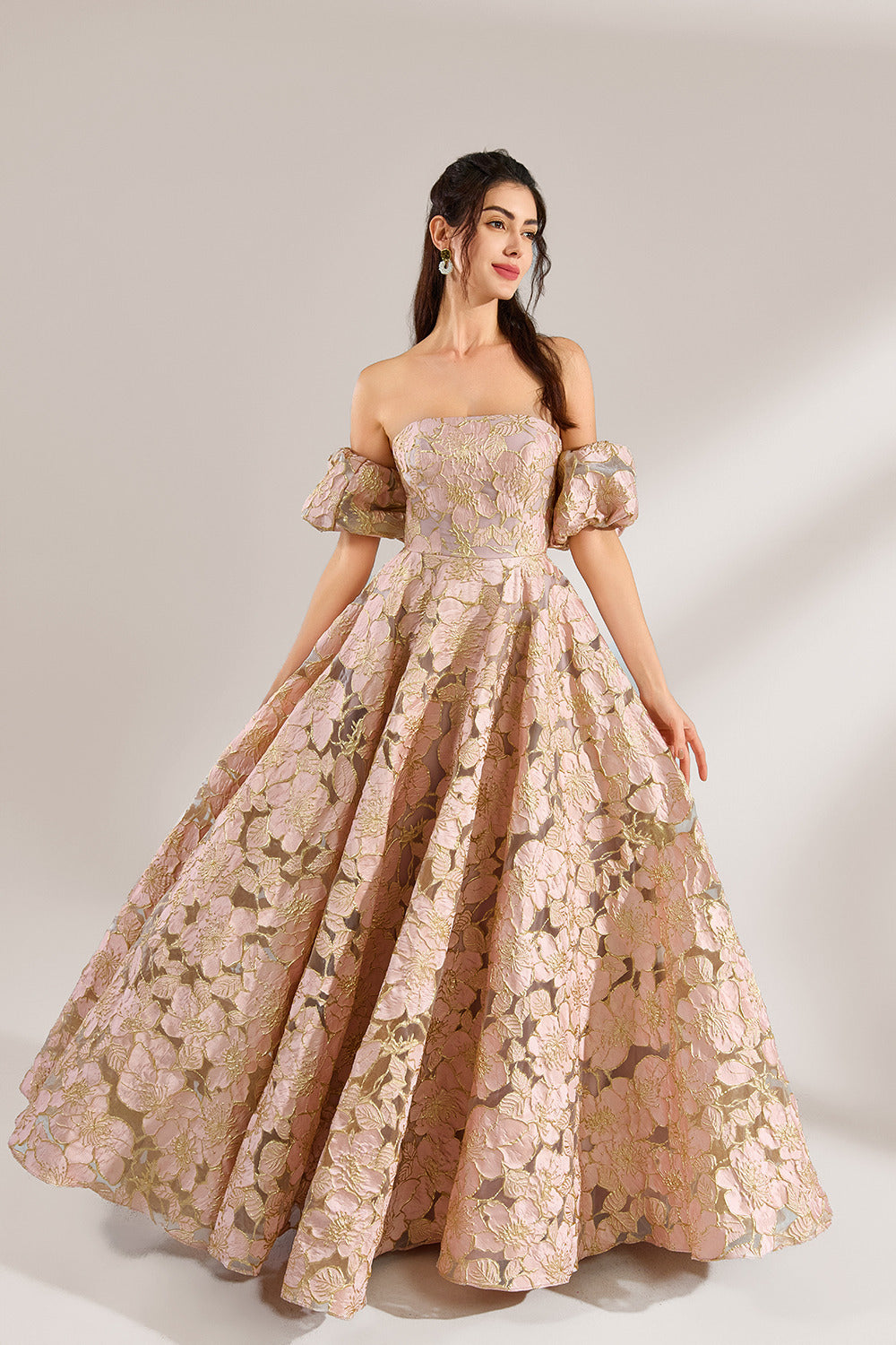 A woman with long, wavy dark hair and a poised stance wears a light - pink, floral - embroidered off - the - shoulder gown. It has puffed sleeves, a fitted bodice, and a voluminous skirt. Paired with understated heels, the outfit radiates elegance, perfect for formal events like galas or weddings.1