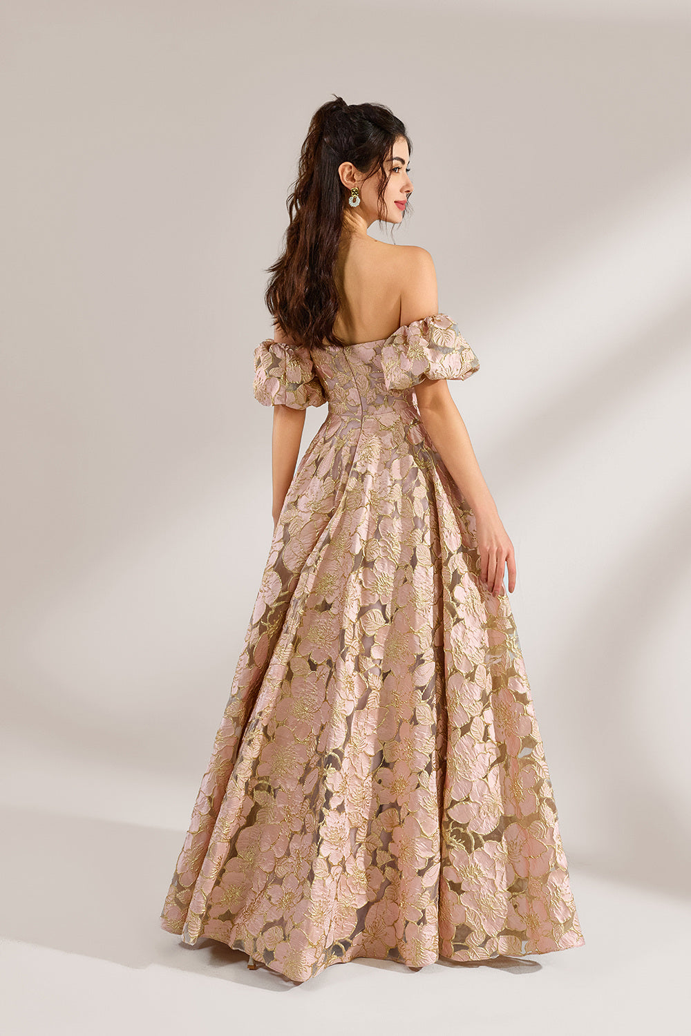 A woman with long, wavy dark hair and a poised stance wears a light - pink, floral - embroidered off - the - shoulder gown. It has puffed sleeves, a fitted bodice, and a voluminous skirt. Paired with understated heels, the outfit radiates elegance, perfect for formal events like galas or weddings.2
