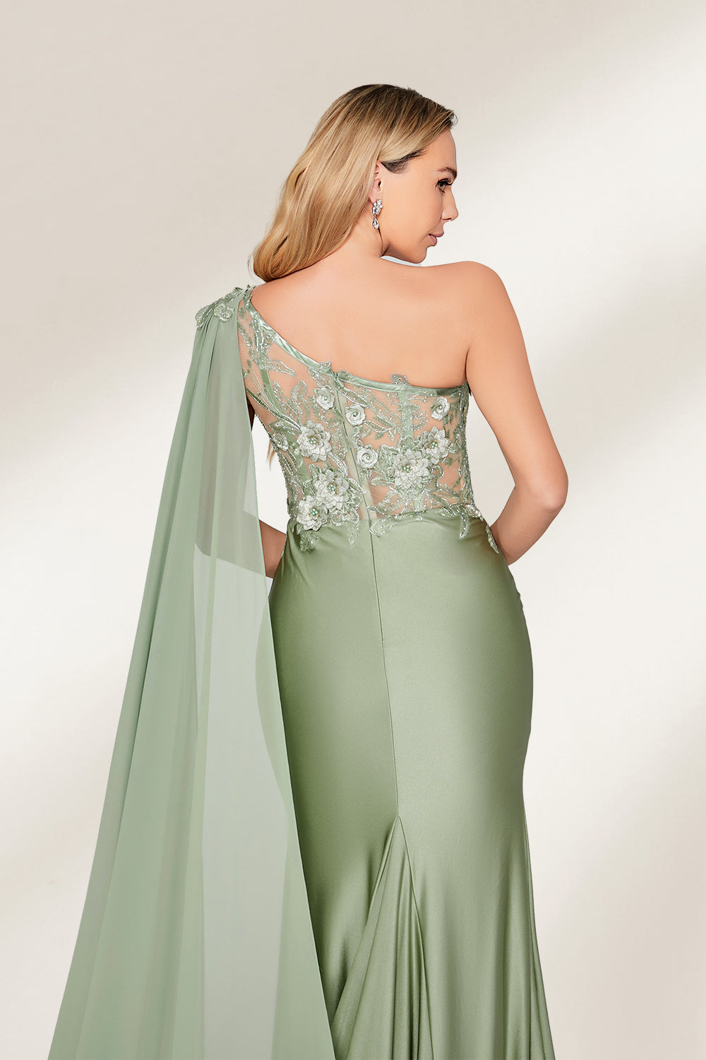 A woman stands against a light background, wearing a sage - green satin gown. It features a one - shoulder design with a flowing cape, floral lace embroidery on the bodice and skirt, and a mermaid silhouette. The elegant details and unique cape create a sophisticated look, perfect for formal events like galas or weddings.6