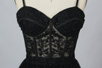 Photograph of a black, lace-trimmed, strapless gown with a sweetheart neckline and a full, flowing tulle skirt. The dress is displayed on a white mannequin with a wooden stand against a plain, light beige background, creating a simple and elegant setting.00
