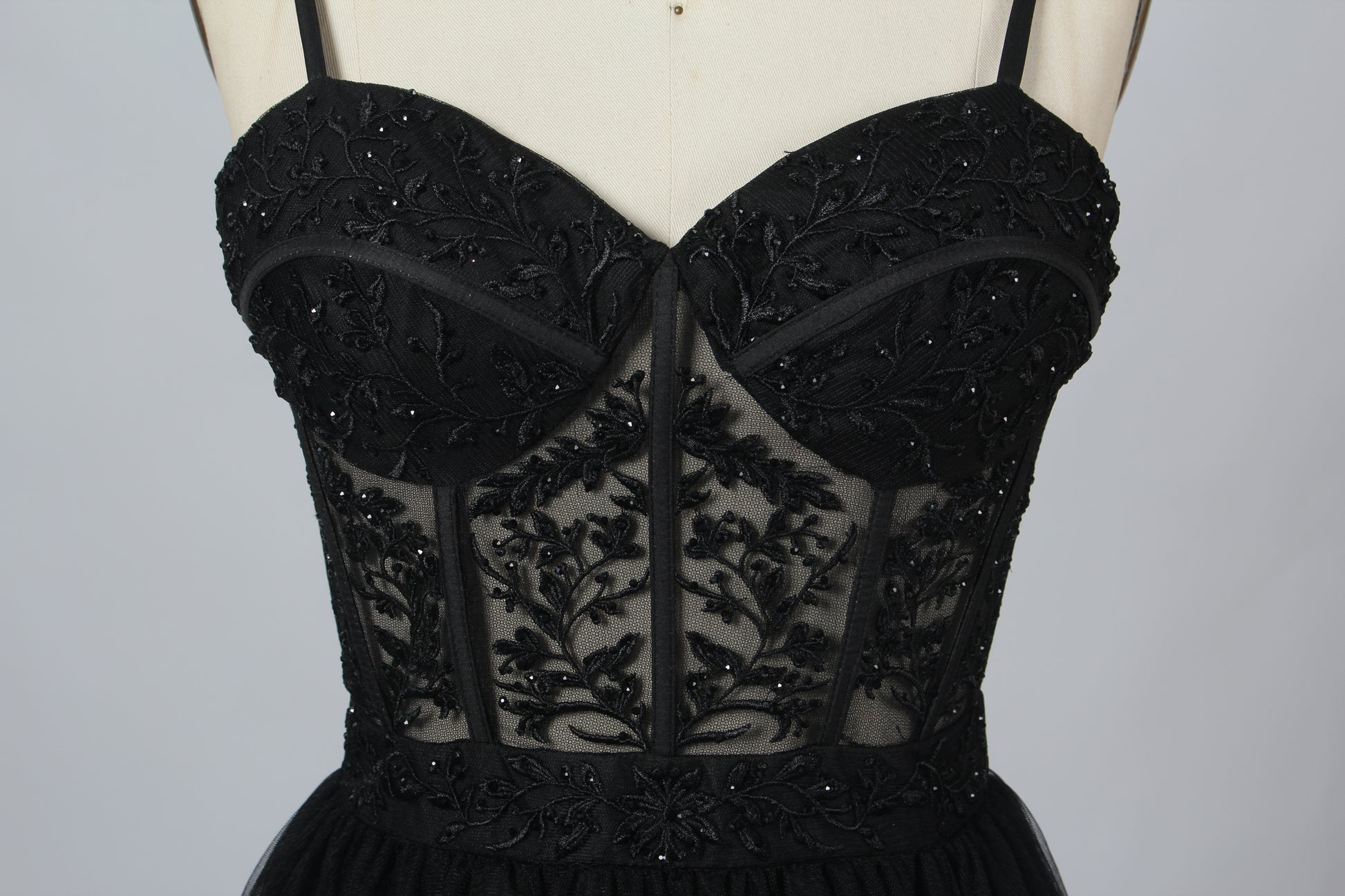Photograph of a black, lace-trimmed, strapless gown with a sweetheart neckline and a full, flowing tulle skirt. The dress is displayed on a white mannequin with a wooden stand against a plain, light beige background, creating a simple and elegant setting.00