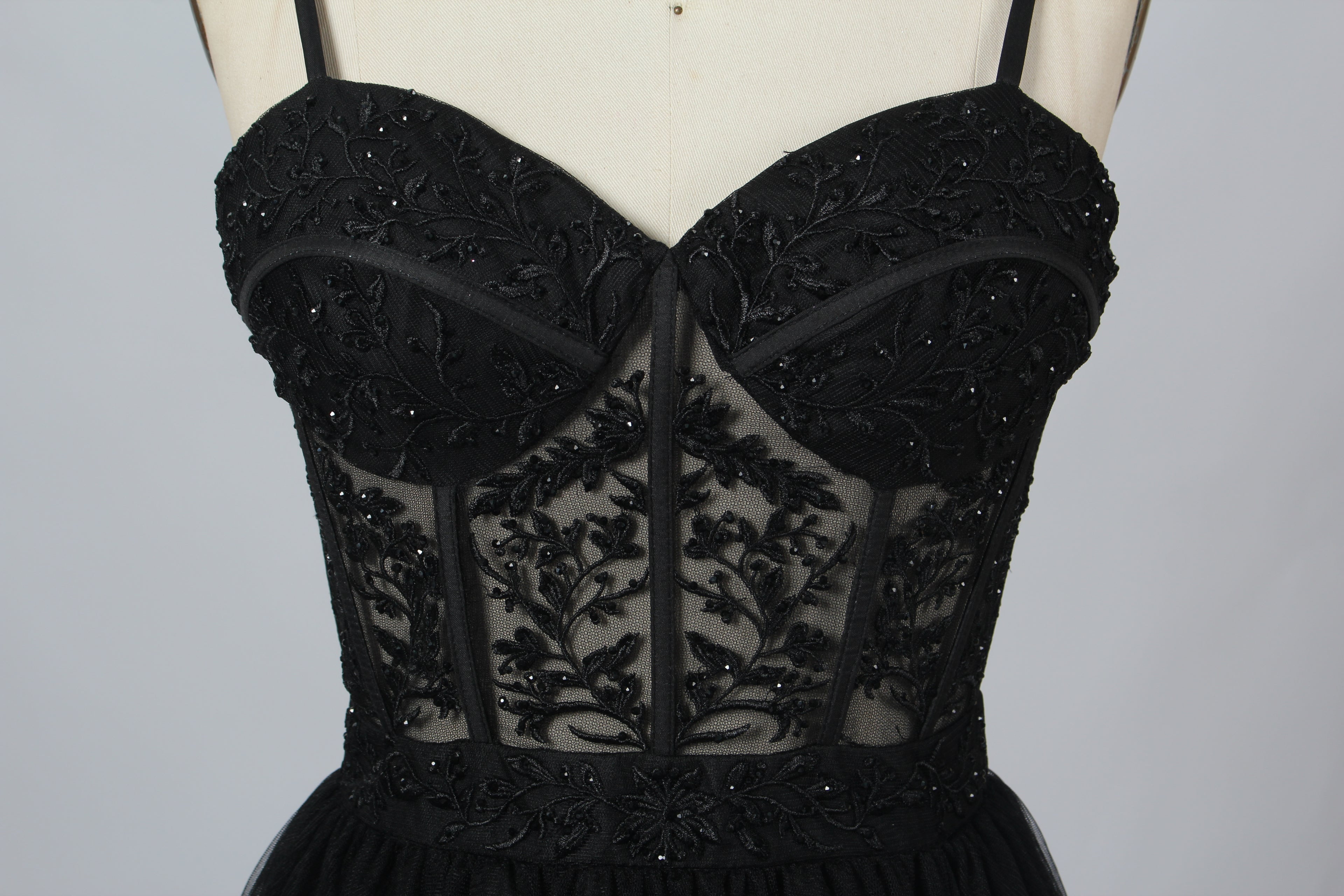 Photograph of a black, lace-trimmed, strapless gown with a sweetheart neckline and a full, flowing tulle skirt. The dress is displayed on a white mannequin with a wooden stand against a plain, light beige background, creating a simple and elegant setting.00