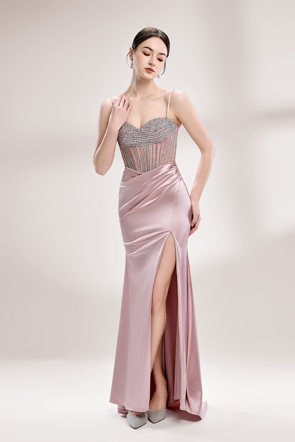 A woman stands against a light background, wearing a pink satin gown with a beaded, sheer bodice. It has thin straps, a ruched skirt, and a high side slit. The contrast of the sparkling beads and smooth satin fabric exudes elegance, perfect for formal galas or evening events, radiating charm.1
