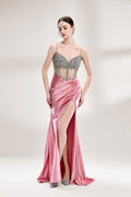 A woman stands against a light - hued background, wearing a pink satin gown with a bejeweled, sheer bodice. It features thin straps, a deep neckline, and a high side slit. Silver - toned heels complement the look, with the sparkling embellishments and elegant silhouette perfect for formal galas.1