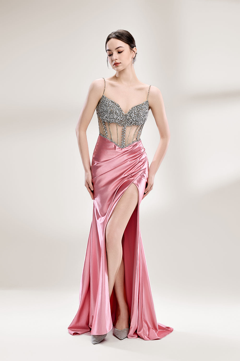 A woman stands against a light - hued background, wearing a pink satin gown with a bejeweled, sheer bodice. It features thin straps, a deep neckline, and a high side slit. Silver - toned heels complement the look, with the sparkling embellishments and elegant silhouette perfect for formal galas.1