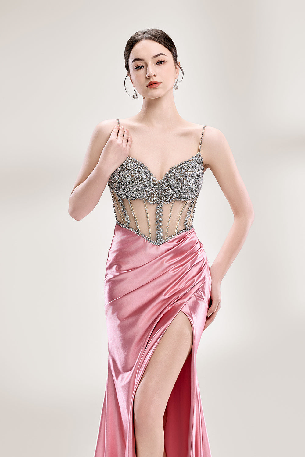 A woman stands against a light - hued background, wearing a pink satin gown with a bejeweled, sheer bodice. It features thin straps, a deep neckline, and a high side slit. Silver - toned heels complement the look, with the sparkling embellishments and elegant silhouette perfect for formal galas.4