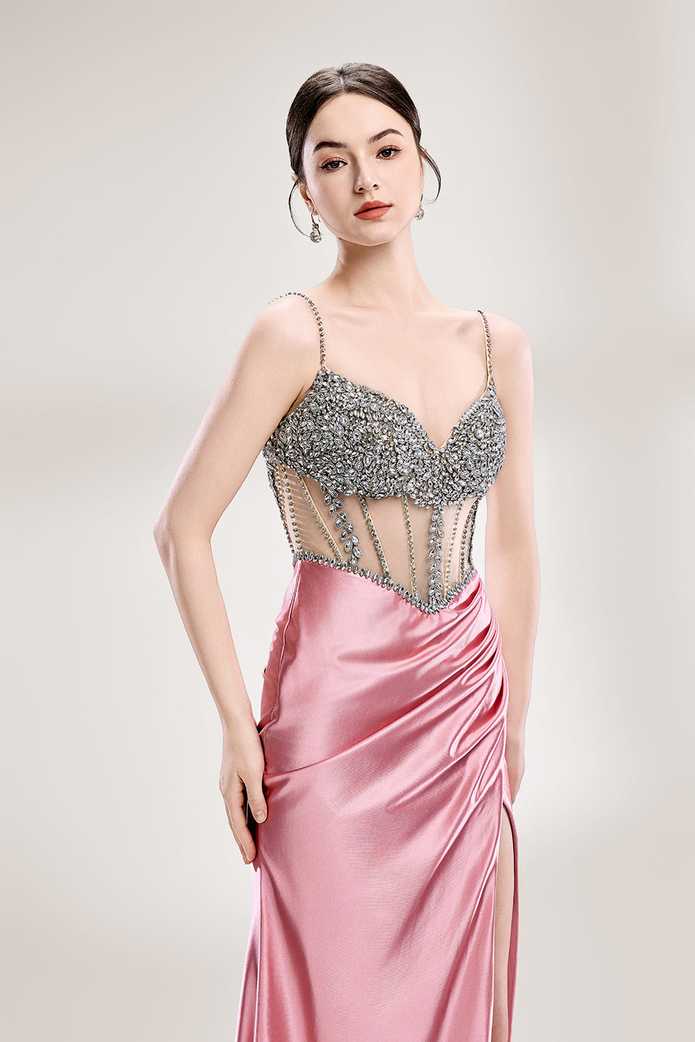 A woman stands against a light - hued background, wearing a pink satin gown with a bejeweled, sheer bodice. It features thin straps, a deep neckline, and a high side slit. Silver - toned heels complement the look, with the sparkling embellishments and elegant silhouette perfect for formal galas.5