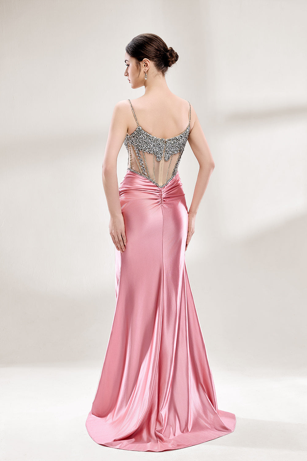 A woman stands against a light - hued background, wearing a pink satin gown with a bejeweled, sheer bodice. It features thin straps, a deep neckline, and a high side slit. Silver - toned heels complement the look, with the sparkling embellishments and elegant silhouette perfect for formal galas.2