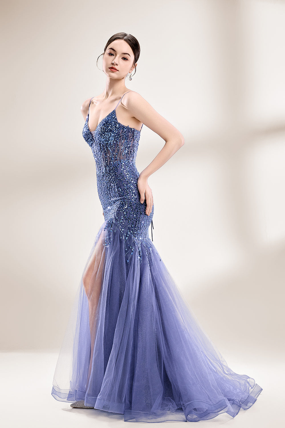 Photograph of a young Asian woman with fair skin and dark hair in a sleek bun, wearing a stunning, floor-length, deep blue evening gown. The gown has a plunging V-neckline, adorned with intricate beading and sequins, and features a sheer overlay with a high slit on one side, revealing a layer of tulle. The background is a soft, neutral gradient, highlighting the gown's elegance. She accessorizes with large, dangling earrings.3