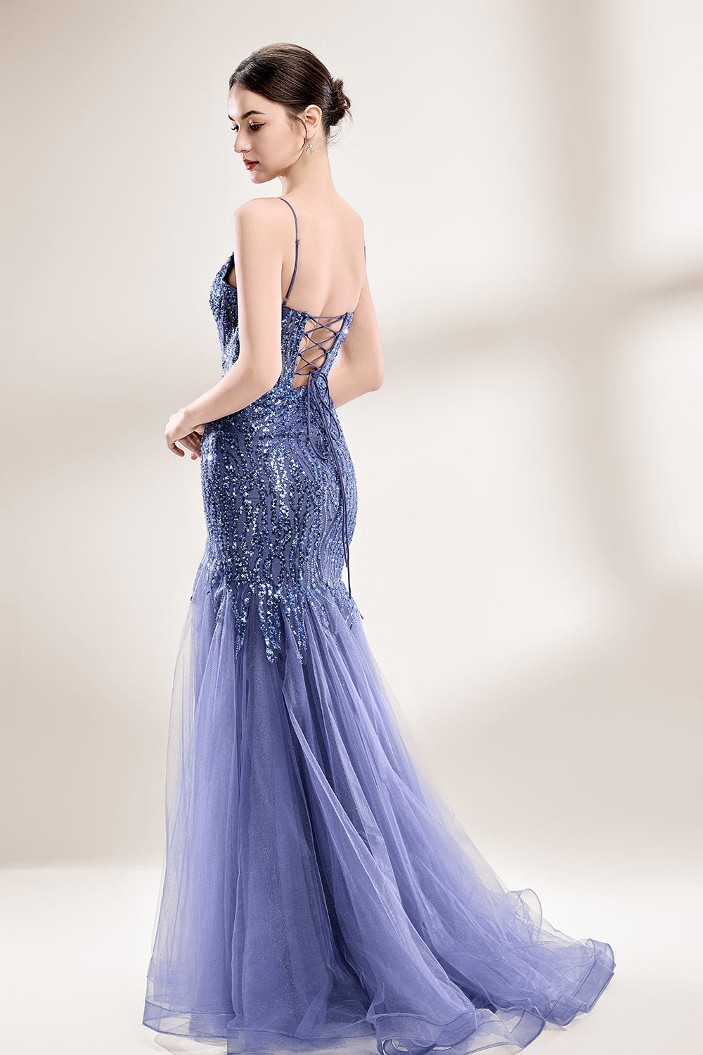 Photograph of a young Asian woman with fair skin and dark hair in a sleek bun, wearing a stunning, floor-length, deep blue evening gown. The gown has a plunging V-neckline, adorned with intricate beading and sequins, and features a sheer overlay with a high slit on one side, revealing a layer of tulle. The background is a soft, neutral gradient, highlighting the gown's elegance. She accessorizes with large, dangling earrings.4
