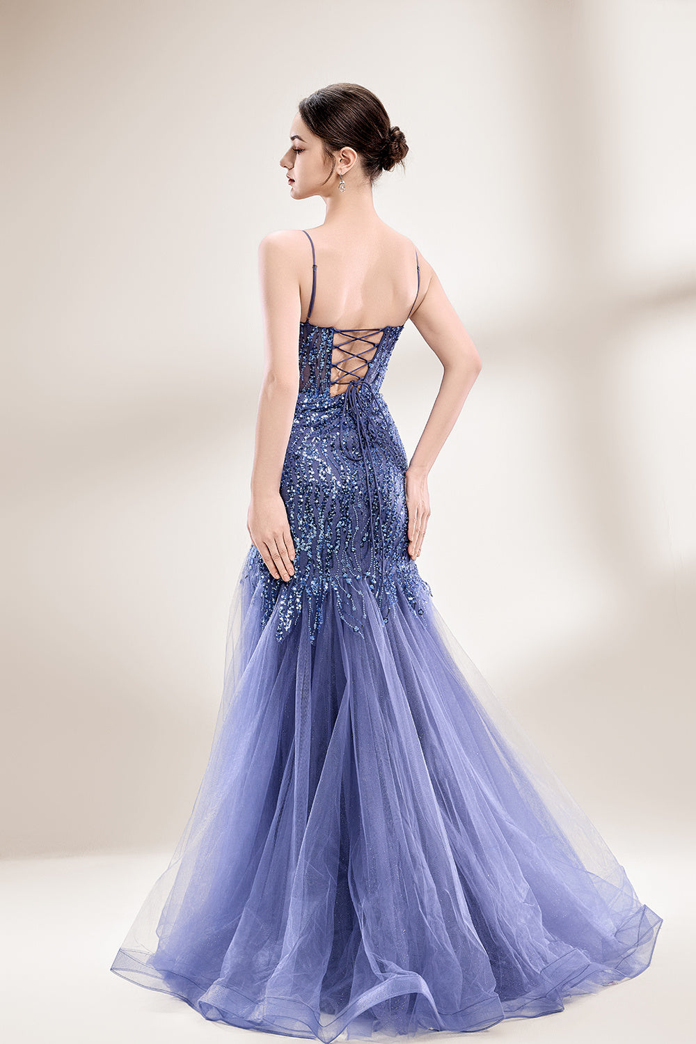Photograph of a young Asian woman with fair skin and dark hair in a sleek bun, wearing a stunning, floor-length, deep blue evening gown. The gown has a plunging V-neckline, adorned with intricate beading and sequins, and features a sheer overlay with a high slit on one side, revealing a layer of tulle. The background is a soft, neutral gradient, highlighting the gown's elegance. She accessorizes with large, dangling earrings.2