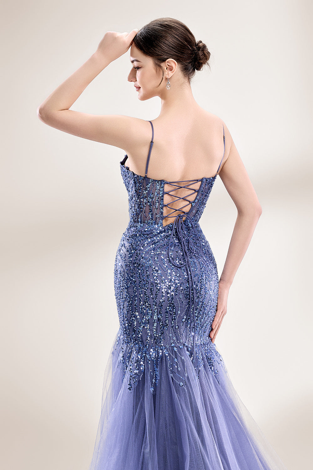 Photograph of a young Asian woman with fair skin and dark hair in a sleek bun, wearing a stunning, floor-length, deep blue evening gown. The gown has a plunging V-neckline, adorned with intricate beading and sequins, and features a sheer overlay with a high slit on one side, revealing a layer of tulle. The background is a soft, neutral gradient, highlighting the gown's elegance. She accessorizes with large, dangling earrings.6