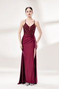 Photograph of a young woman standing against a plain, light gray background. She has fair skin, dark brown hair styled in a sleek bun, and is wearing a burgundy evening gown with intricate lace detailing on the bodice and a high slit on the right. The gown has thin spaghetti straps and a deep V-neckline. She is accessorized with silver high heels and large hoop earrings. The overall style is elegant and sophisticated, suitable for a formal event.1