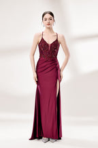 Photograph of a young woman standing against a plain, light gray background. She has fair skin, dark brown hair styled in a sleek bun, and is wearing a burgundy evening gown with intricate lace detailing on the bodice and a high slit on the right. The gown has thin spaghetti straps and a deep V-neckline. She is accessorized with silver high heels and large hoop earrings. The overall style is elegant and sophisticated, suitable for a formal event.1