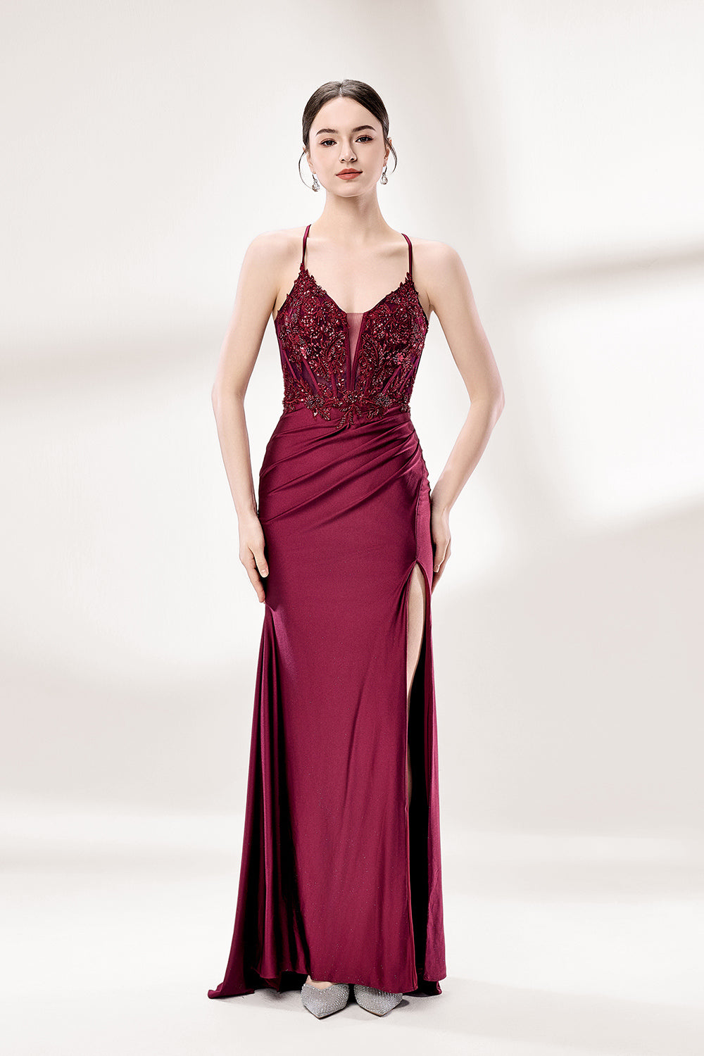 Photograph of a young woman standing against a plain, light gray background. She has fair skin, dark brown hair styled in a sleek bun, and is wearing a burgundy evening gown with intricate lace detailing on the bodice and a high slit on the right. The gown has thin spaghetti straps and a deep V-neckline. She is accessorized with silver high heels and large hoop earrings. The overall style is elegant and sophisticated, suitable for a formal event.1