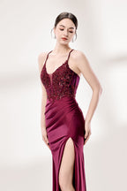 Photograph of a young woman standing against a plain, light gray background. She has fair skin, dark brown hair styled in a sleek bun, and is wearing a burgundy evening gown with intricate lace detailing on the bodice and a high slit on the right. The gown has thin spaghetti straps and a deep V-neckline. She is accessorized with silver high heels and large hoop earrings. The overall style is elegant and sophisticated, suitable for a formal event.6