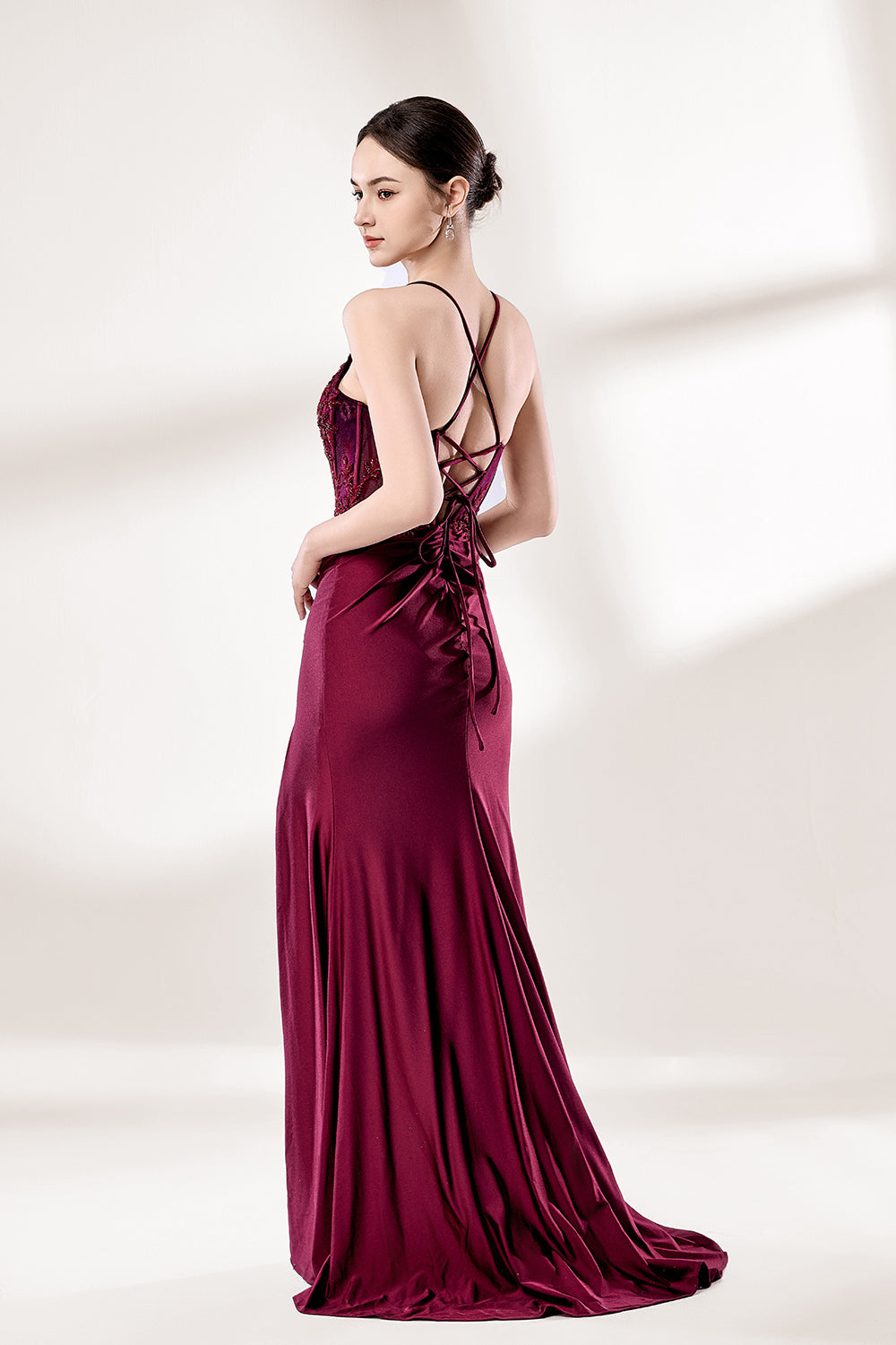 Photograph of a young woman standing against a plain, light gray background. She has fair skin, dark brown hair styled in a sleek bun, and is wearing a burgundy evening gown with intricate lace detailing on the bodice and a high slit on the right. The gown has thin spaghetti straps and a deep V-neckline. She is accessorized with silver high heels and large hoop earrings. The overall style is elegant and sophisticated, suitable for a formal event.4