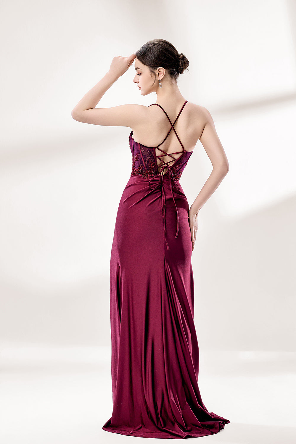 Photograph of a young woman standing against a plain, light gray background. She has fair skin, dark brown hair styled in a sleek bun, and is wearing a burgundy evening gown with intricate lace detailing on the bodice and a high slit on the right. The gown has thin spaghetti straps and a deep V-neckline. She is accessorized with silver high heels and large hoop earrings. The overall style is elegant and sophisticated, suitable for a formal event.2