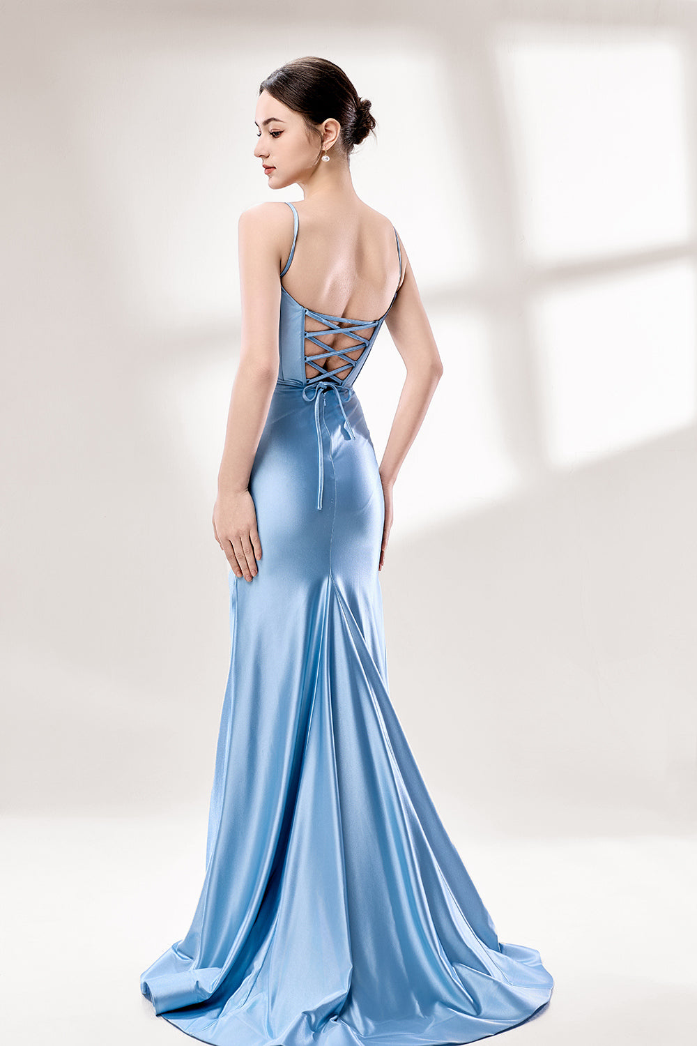 Photograph of a slender, fair-skinned woman with dark hair in an elegant updo, wearing a stunning, floor-length, light blue satin gown with a plunging neckline and thin spaghetti straps. The dress features a fitted bodice and a flowing skirt. She stands against a plain, light gray background, with soft lighting emphasizing the gown's sheen.2