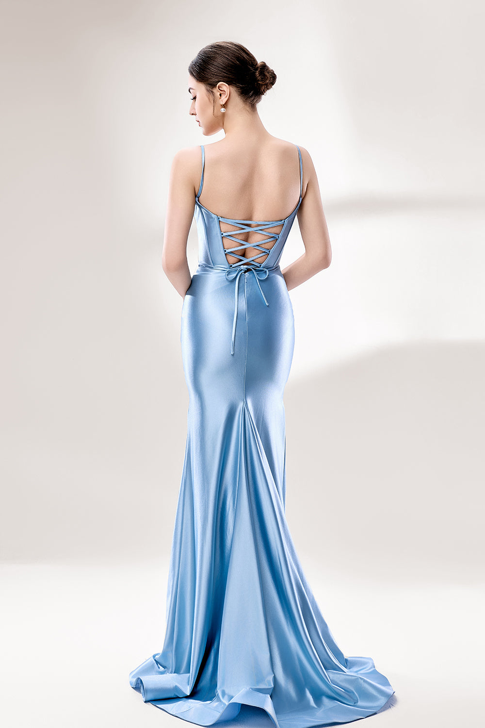 Photograph of a slender, fair-skinned woman with dark hair in an elegant updo, wearing a stunning, floor-length, light blue satin gown with a plunging neckline and thin spaghetti straps. The dress features a fitted bodice and a flowing skirt. She stands against a plain, light gray background, with soft lighting emphasizing the gown's sheen.5
