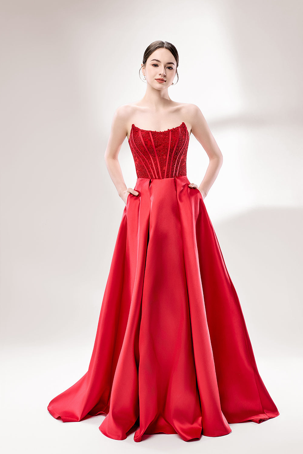 Against a light - hued background, a woman wears a red satin gown with a strapless, lace - detailed bodice and pockets. It has a voluminous skirt, combining romance and functionality. The vibrant red color and elegant design radiate confidence, perfect for formal events like weddings or award ceremonies.1