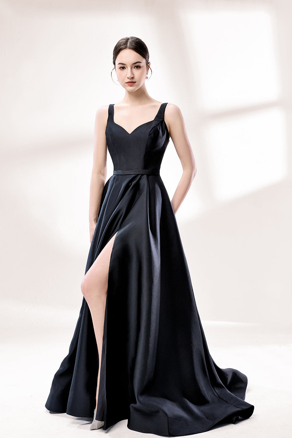 A woman stands against a light background, wearing a black satin gown. It features thin straps, a V - neckline, and a high side slit. The smooth satin fabric and elegant silhouette exude sophistication, perfect for formal events like galas or charity balls, radiating timeless charm.1