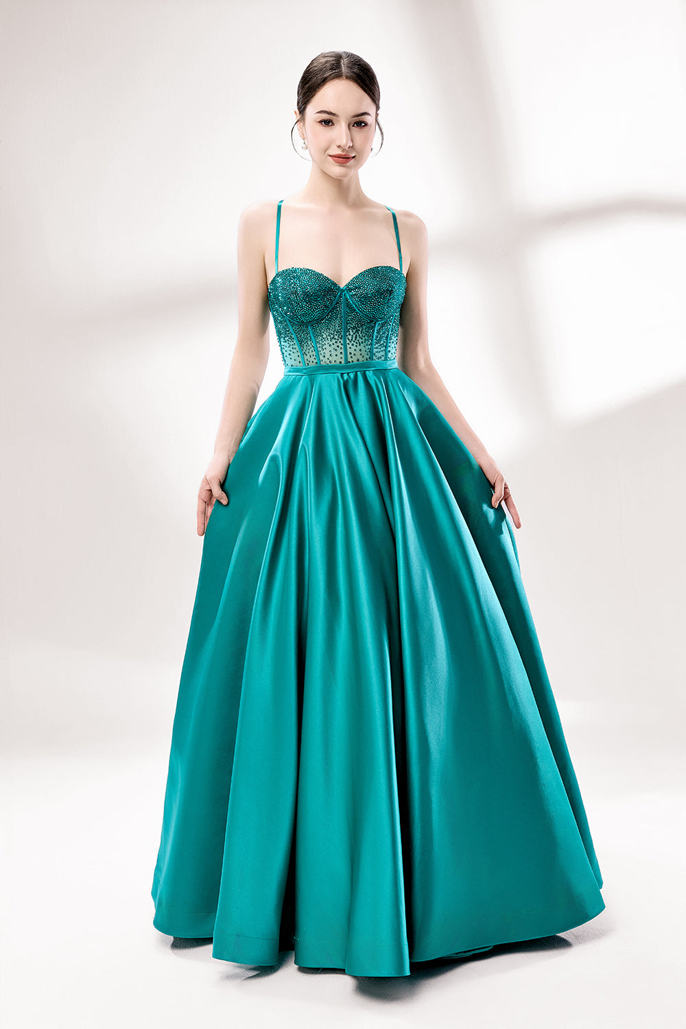 A woman stands against a light background, wearing a teal satin gown. It has thin straps, a sweetheart neckline, and a voluminous skirt. The bodice features sheer fabric with bead embellishments, combining elegance and romance. Perfect for formal events like weddings, it exudes charm.3