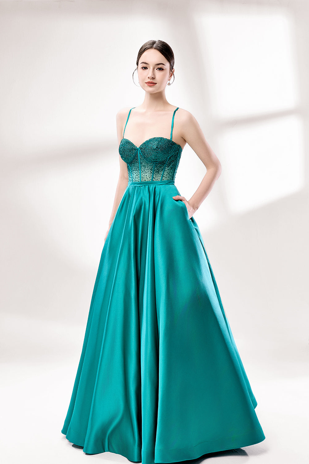 A woman stands against a light background, wearing a teal satin gown. It has thin straps, a sweetheart neckline, and a voluminous skirt. The bodice features sheer fabric with bead embellishments, combining elegance and romance. Perfect for formal events like weddings, it exudes charm.1