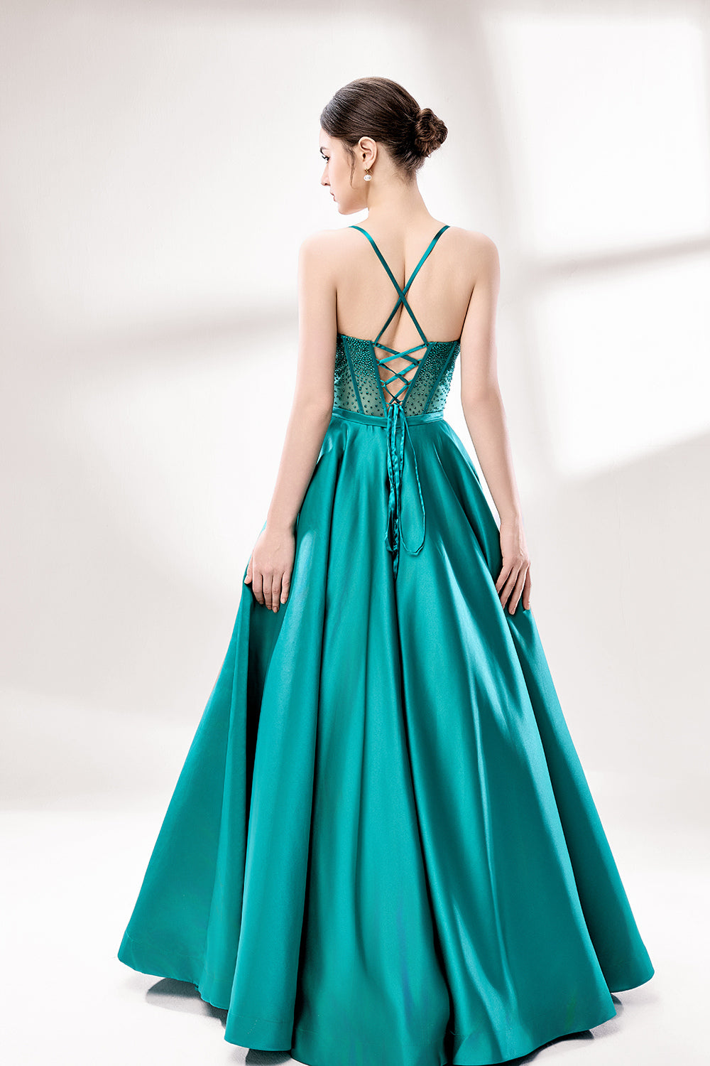 A woman stands against a light background, wearing a teal satin gown. It has thin straps, a sweetheart neckline, and a voluminous skirt. The bodice features sheer fabric with bead embellishments, combining elegance and romance. Perfect for formal events like weddings, it exudes charm.2