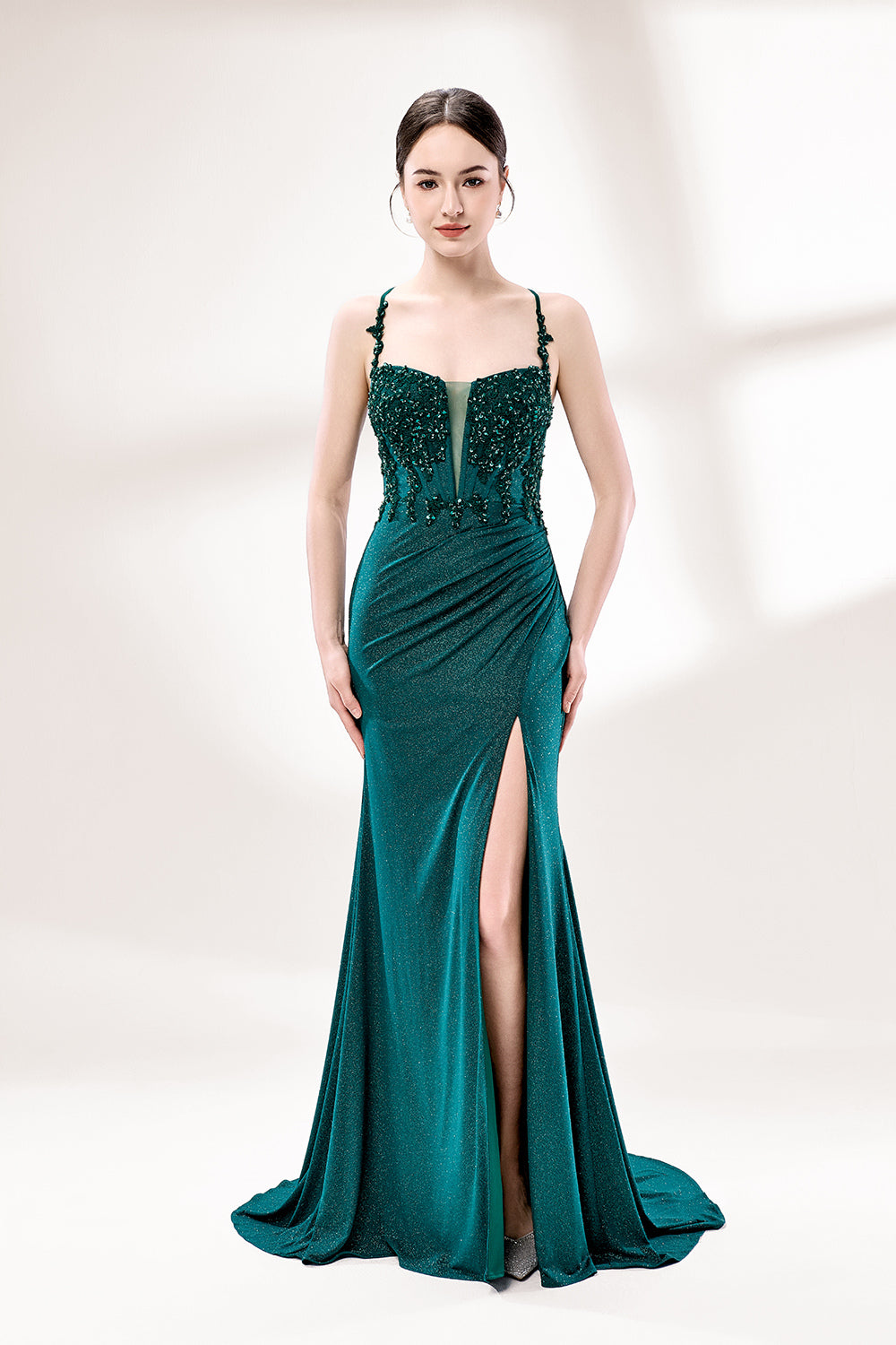 Photograph of a young Caucasian woman with fair skin and dark brown hair styled in an elegant updo. She is wearing a stunning, floor-length, teal evening gown with a deep V-neckline adorned with intricate beadwork. The gown features a high slit on the left, showcasing her slender legs. The fabric has a smooth, shimmering texture. The background is a soft, white gradient, creating a minimalist and elegant atmosphere. The overall style is sophisticated and glamorous, suitable for formal events.1