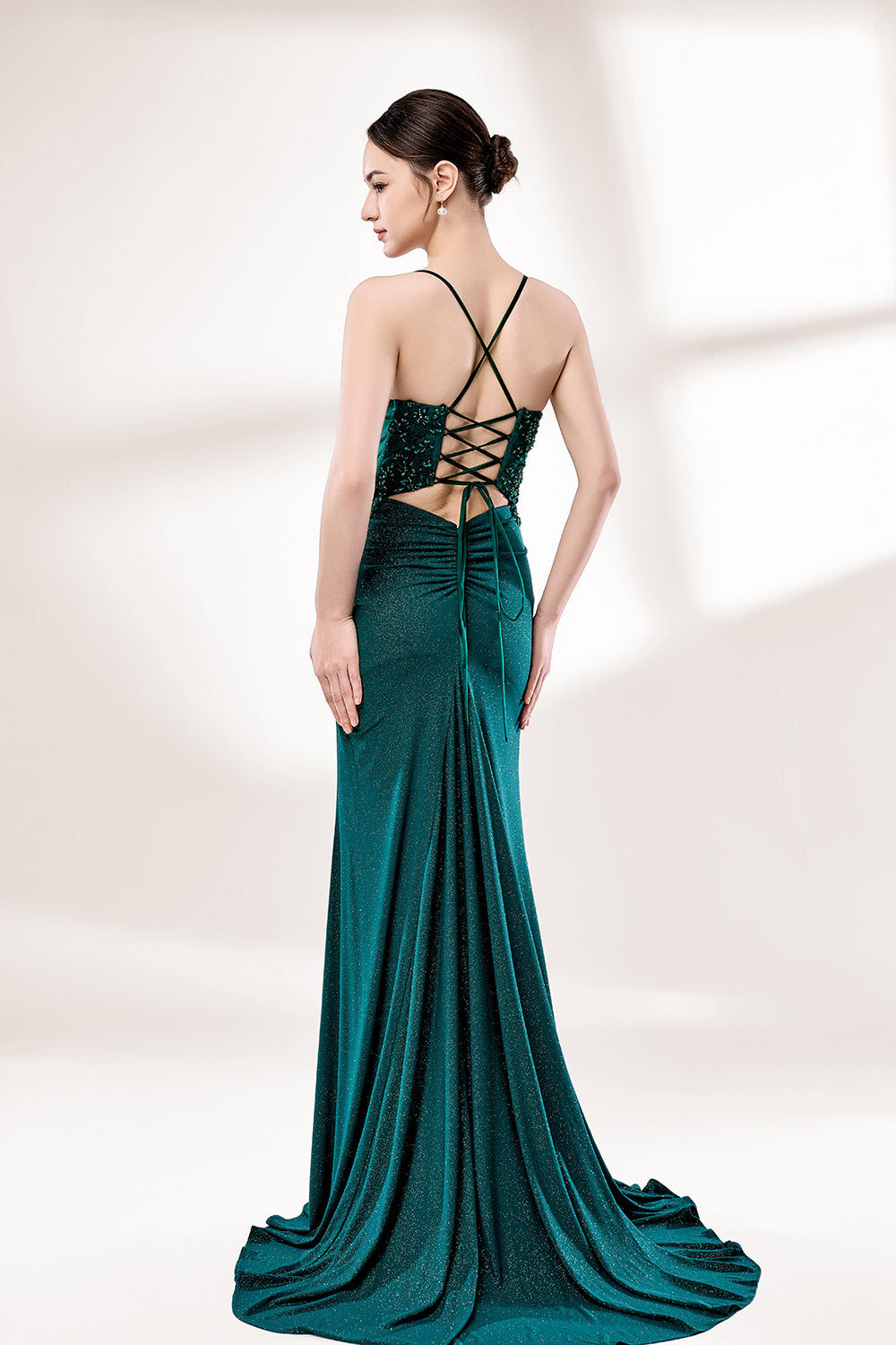 Photograph of a young Caucasian woman with fair skin and dark brown hair styled in an elegant updo. She is wearing a stunning, floor-length, teal evening gown with a deep V-neckline adorned with intricate beadwork. The gown features a high slit on the left, showcasing her slender legs. The fabric has a smooth, shimmering texture. The background is a soft, white gradient, creating a minimalist and elegant atmosphere. The overall style is sophisticated and glamorous, suitable for formal events.2