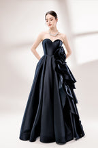 Bathed in soft light, a woman dazzles in a black strapless ball gown. It features a sweetheart neckline, a large ruffled floral detail on the side, and a voluminous skirt. The elegant design creates a sophisticated and glamorous look, perfect for formal events like galas or weddings.1
