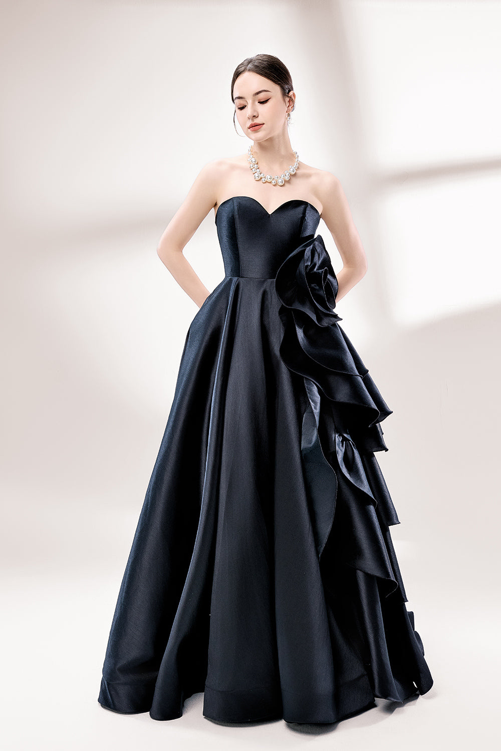 Bathed in soft light, a woman dazzles in a black strapless ball gown. It features a sweetheart neckline, a large ruffled floral detail on the side, and a voluminous skirt. The elegant design creates a sophisticated and glamorous look, perfect for formal events like galas or weddings.1
