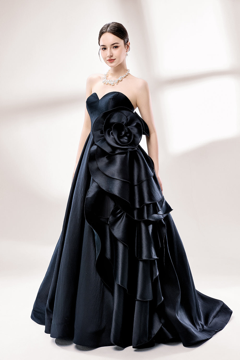 Bathed in soft light, a woman dazzles in a black strapless ball gown. It features a sweetheart neckline, a large ruffled floral detail on the side, and a voluminous skirt. The elegant design creates a sophisticated and glamorous look, perfect for formal events like galas or weddings.2