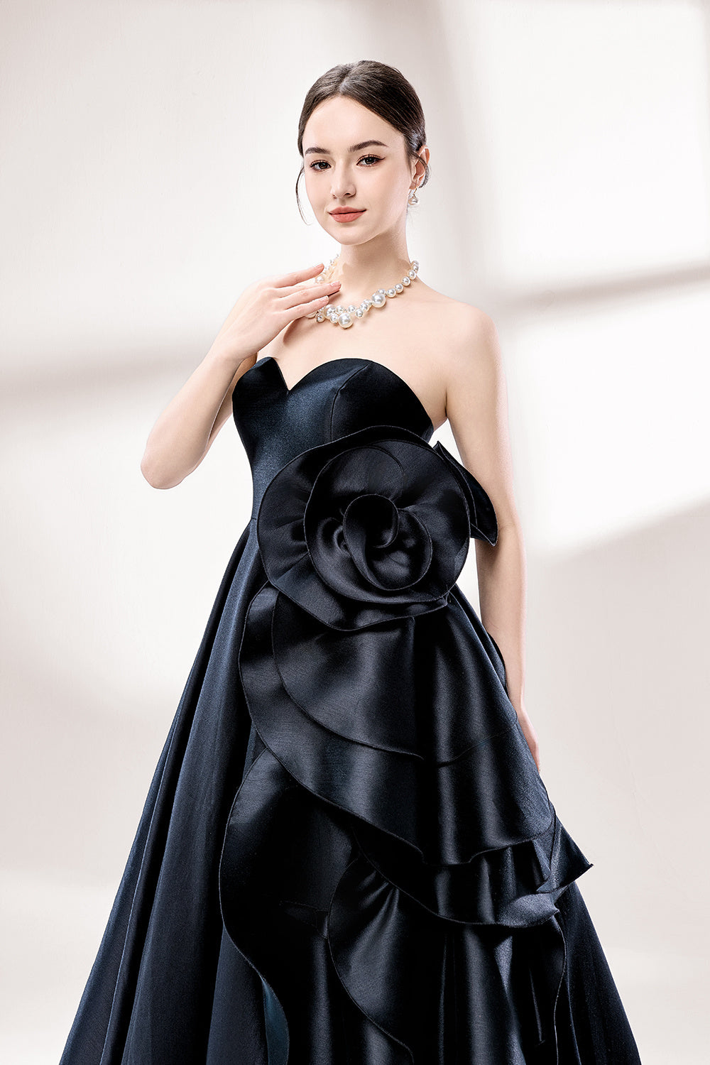 Bathed in soft light, a woman dazzles in a black strapless ball gown. It features a sweetheart neckline, a large ruffled floral detail on the side, and a voluminous skirt. The elegant design creates a sophisticated and glamorous look, perfect for formal events like galas or weddings.3