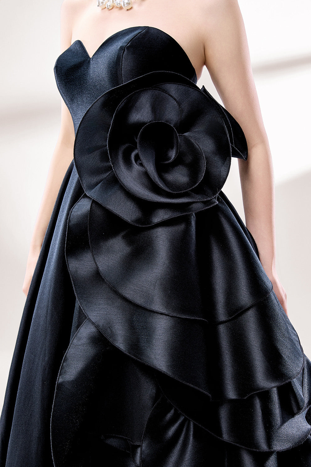 Bathed in soft light, a woman dazzles in a black strapless ball gown. It features a sweetheart neckline, a large ruffled floral detail on the side, and a voluminous skirt. The elegant design creates a sophisticated and glamorous look, perfect for formal events like galas or weddings.4