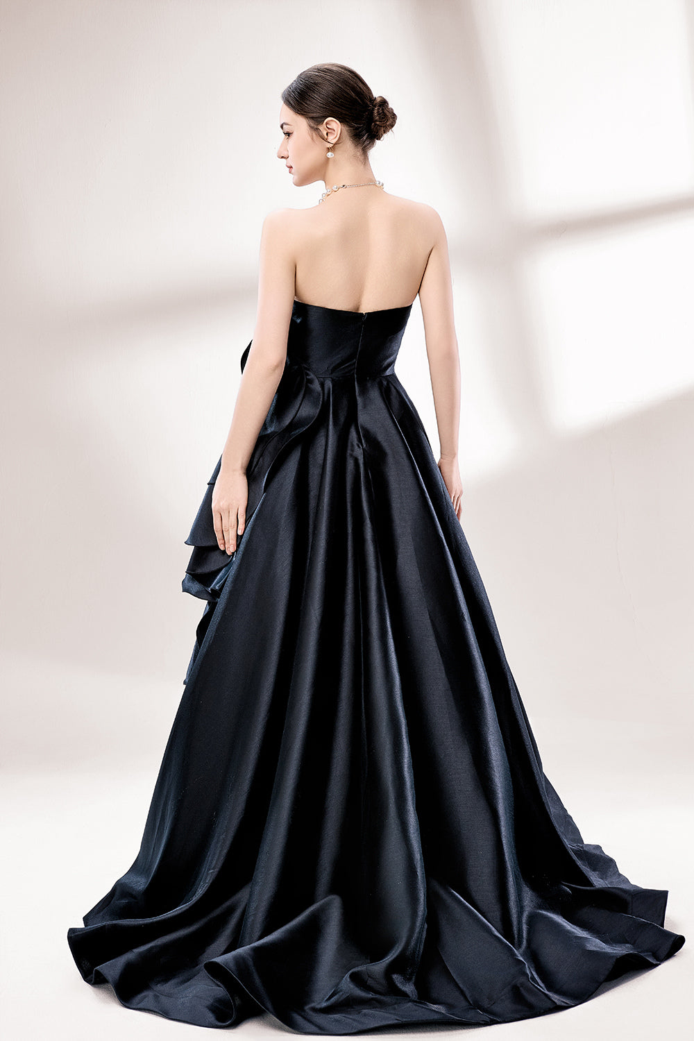 Bathed in soft light, a woman dazzles in a black strapless ball gown. It features a sweetheart neckline, a large ruffled floral detail on the side, and a voluminous skirt. The elegant design creates a sophisticated and glamorous look, perfect for formal events like galas or weddings.5