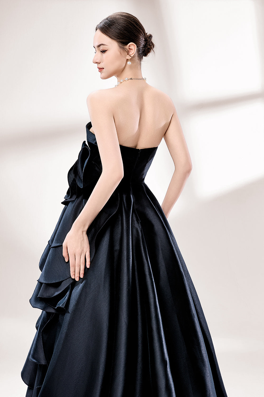 Bathed in soft light, a woman dazzles in a black strapless ball gown. It features a sweetheart neckline, a large ruffled floral detail on the side, and a voluminous skirt. The elegant design creates a sophisticated and glamorous look, perfect for formal events like galas or weddings.6