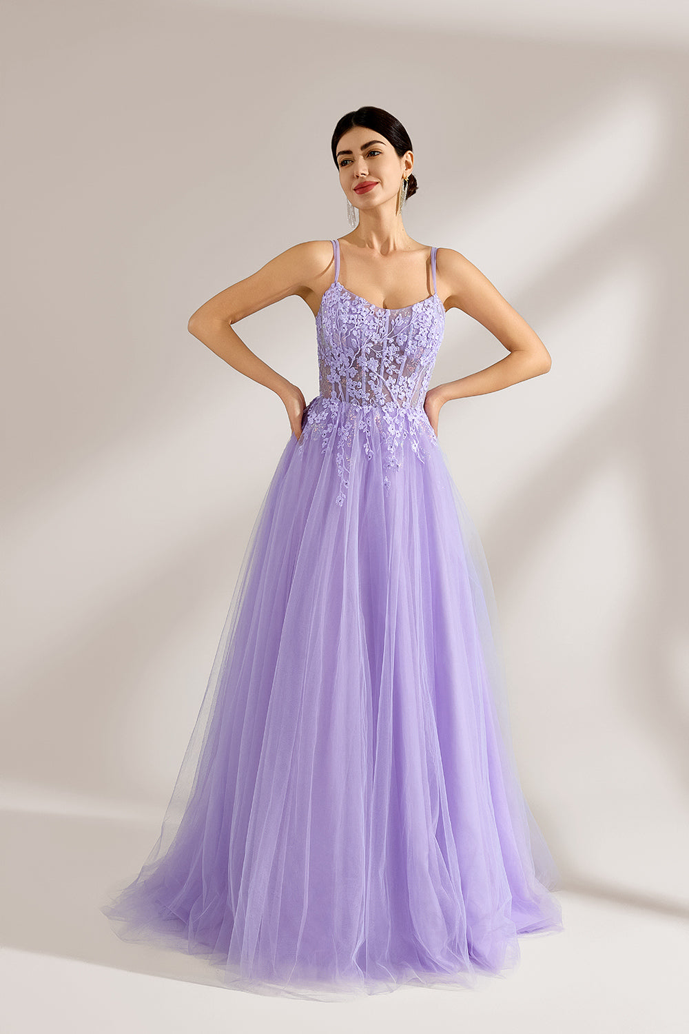 Set against a light backdrop, a woman wears a lavender tulle gown. It has thin straps, a sweetheart neckline with intricate lace embroidery on the bodice, and a flowing tulle skirt. The graceful design gives off a romantic and ethereal vibe, ideal for formal occasions such as proms or weddings.1