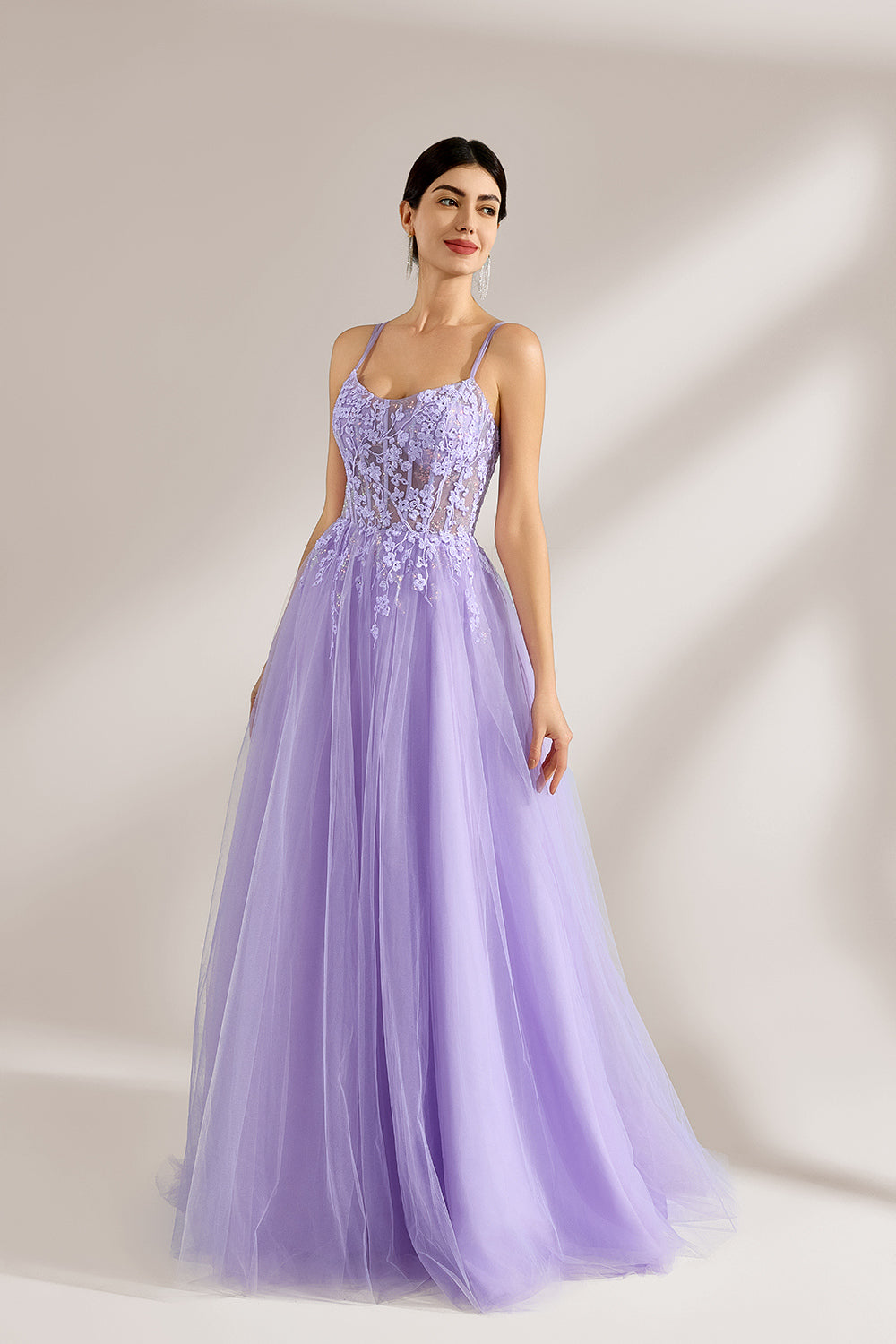 Set against a light backdrop, a woman wears a lavender tulle gown. It has thin straps, a sweetheart neckline with intricate lace embroidery on the bodice, and a flowing tulle skirt. The graceful design gives off a romantic and ethereal vibe, ideal for formal occasions such as proms or weddings.3