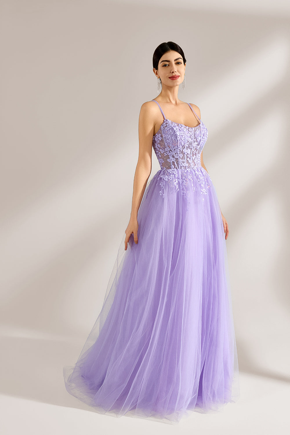 Set against a light backdrop, a woman wears a lavender tulle gown. It has thin straps, a sweetheart neckline with intricate lace embroidery on the bodice, and a flowing tulle skirt. The graceful design gives off a romantic and ethereal vibe, ideal for formal occasions such as proms or weddings.4