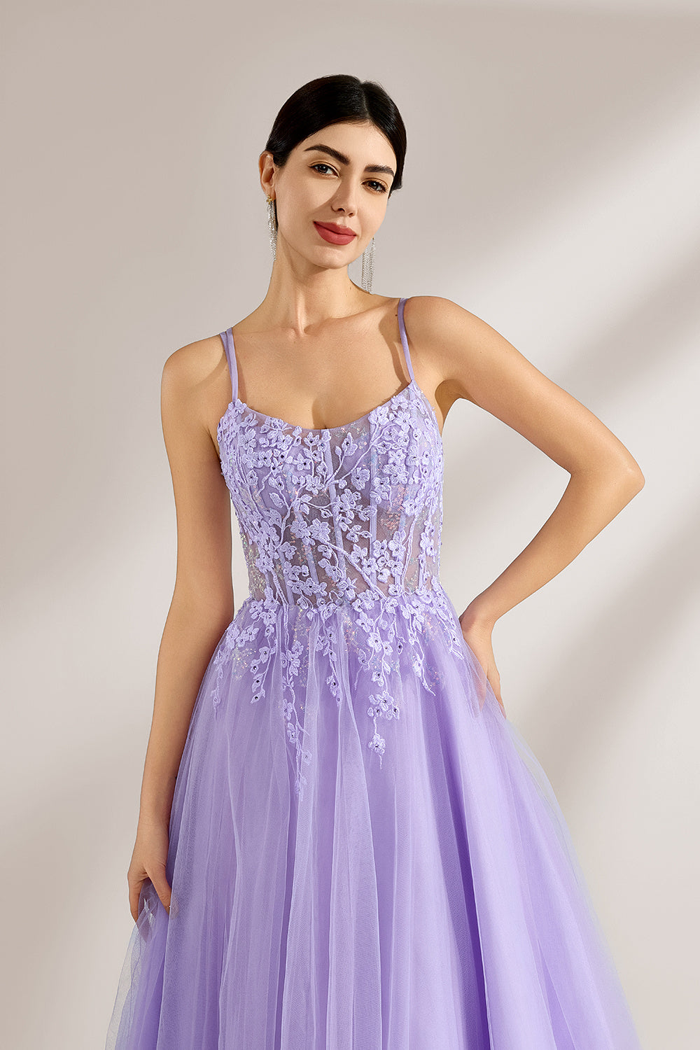Set against a light backdrop, a woman wears a lavender tulle gown. It has thin straps, a sweetheart neckline with intricate lace embroidery on the bodice, and a flowing tulle skirt. The graceful design gives off a romantic and ethereal vibe, ideal for formal occasions such as proms or weddings.5
