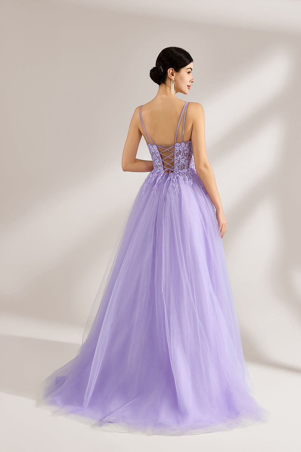 Set against a light backdrop, a woman wears a lavender tulle gown. It has thin straps, a sweetheart neckline with intricate lace embroidery on the bodice, and a flowing tulle skirt. The graceful design gives off a romantic and ethereal vibe, ideal for formal occasions such as proms or weddings.2