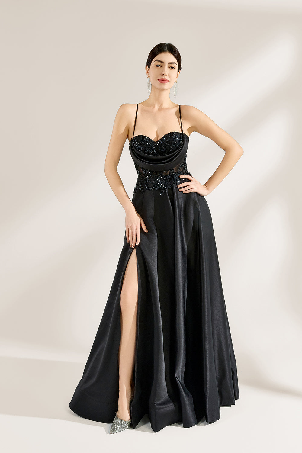 A woman stands against a light background, wearing a black satin gown. The bodice features thin straps, a draped neckline, and beaded embroidery. A high side slit adds allure, combining elegance and sexiness. Perfect for formal events like galas or evening parties, exuding sophistication.1