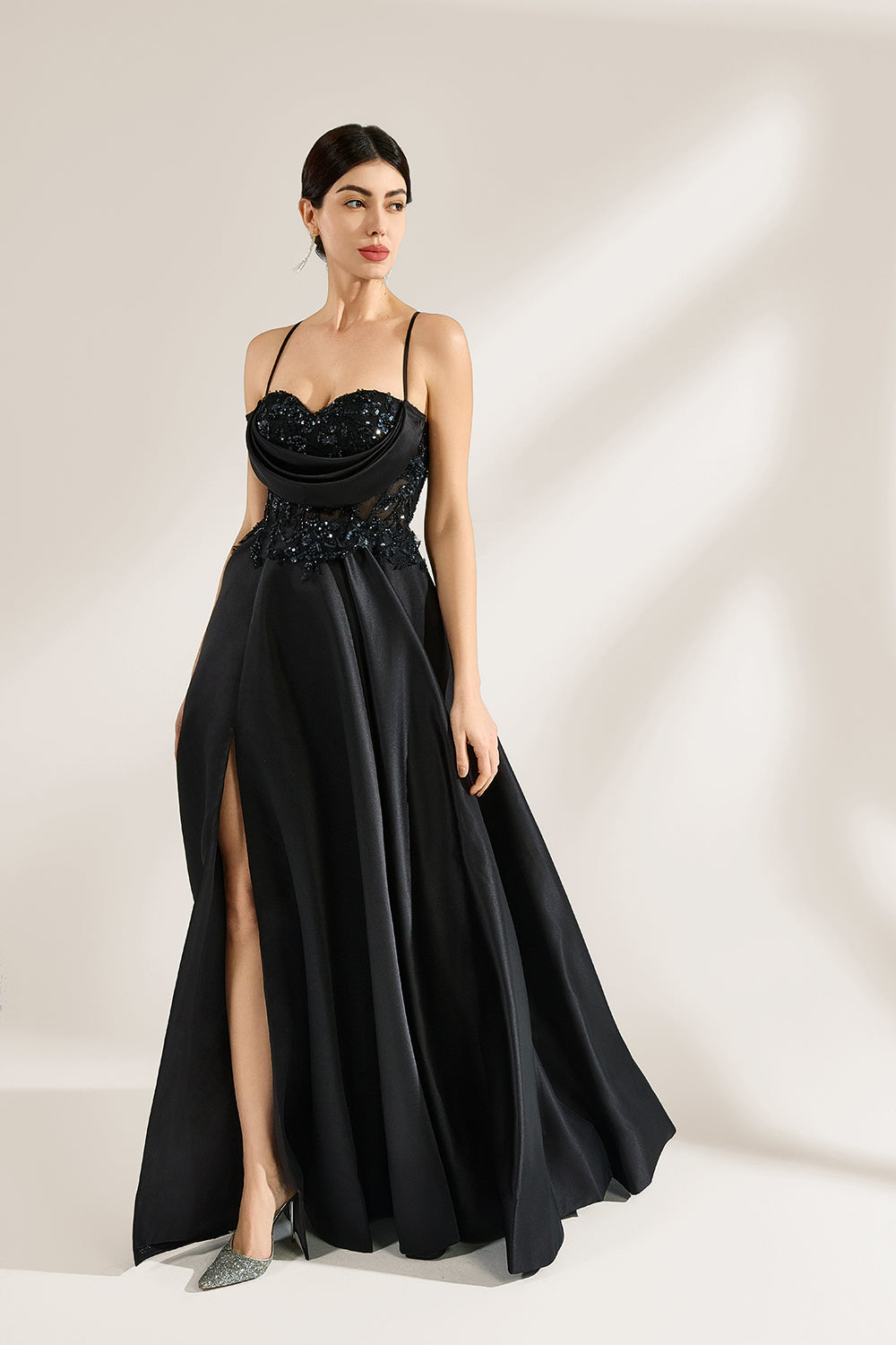 A woman stands against a light background, wearing a black satin gown. The bodice features thin straps, a draped neckline, and beaded embroidery. A high side slit adds allure, combining elegance and sexiness. Perfect for formal events like galas or evening parties, exuding sophistication.3