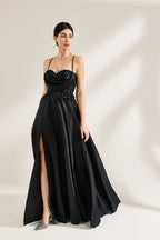 A woman stands against a light background, wearing a black satin gown. The bodice features thin straps, a draped neckline, and beaded embroidery. A high side slit adds allure, combining elegance and sexiness. Perfect for formal events like galas or evening parties, exuding sophistication.3