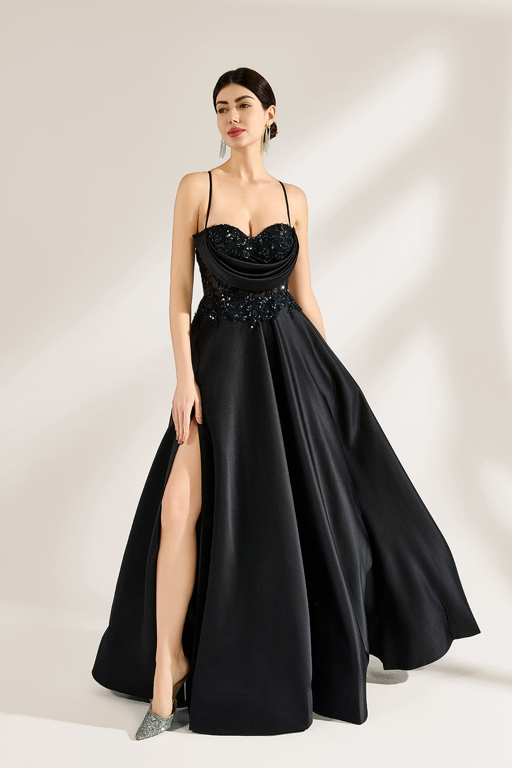 A woman stands against a light background, wearing a black satin gown. The bodice features thin straps, a draped neckline, and beaded embroidery. A high side slit adds allure, combining elegance and sexiness. Perfect for formal events like galas or evening parties, exuding sophistication.4