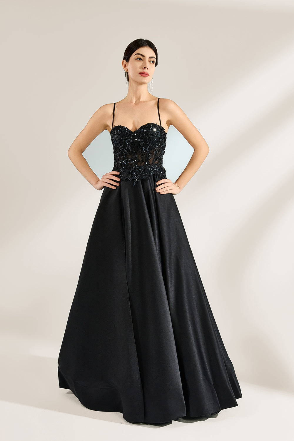 A woman stands against a light background, wearing a black satin gown. The bodice features thin straps, a draped neckline, and beaded embroidery. A high side slit adds allure, combining elegance and sexiness. Perfect for formal events like galas or evening parties, exuding sophistication.5
