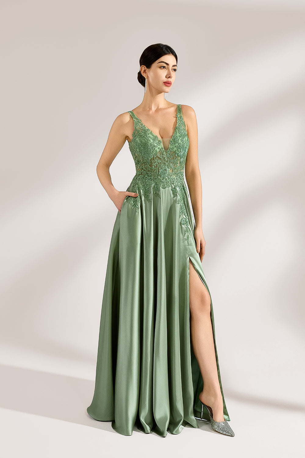 A woman with an elegant updo and a poised stance wears a sage - green satin gown. It has thin straps, a lace - detailed, V - neck bodice, and a high side slit. Paired with silver - tone heels, the outfit radiates grace, perfect for formal events like galas or weddings.2